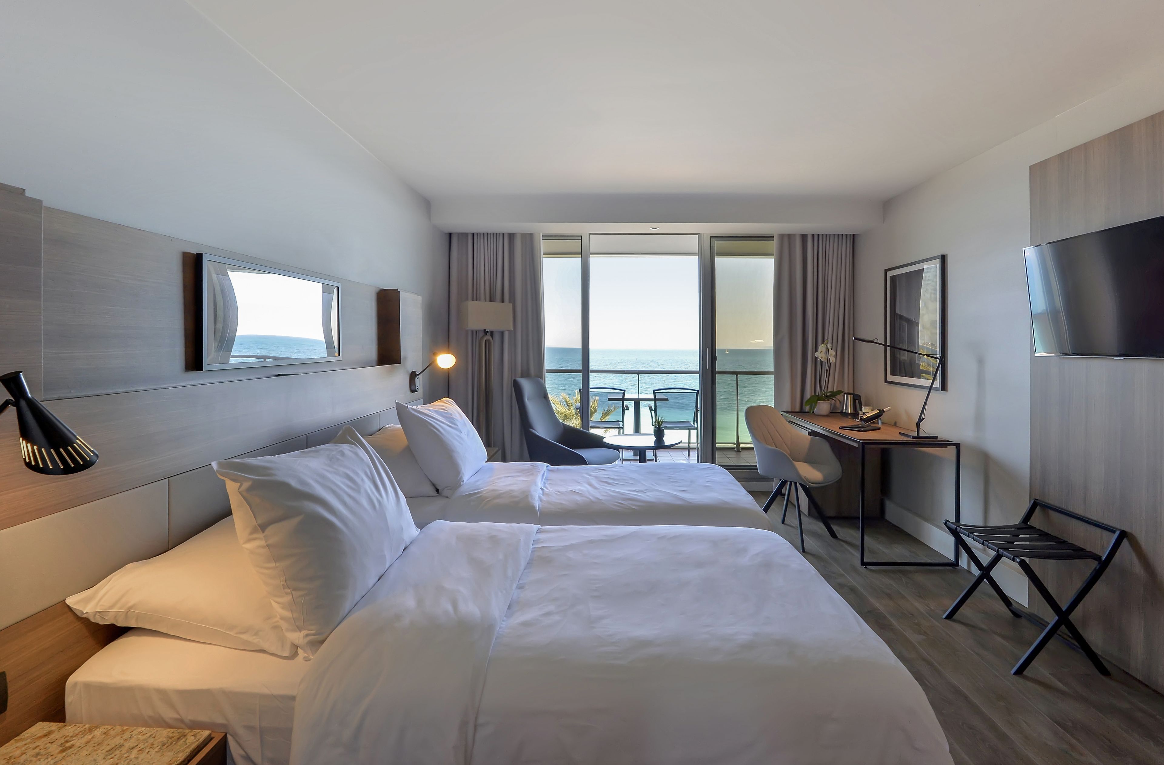 A hotel room with two beds, a desk, a chair, a TV, and a balcony with a view of the sea.