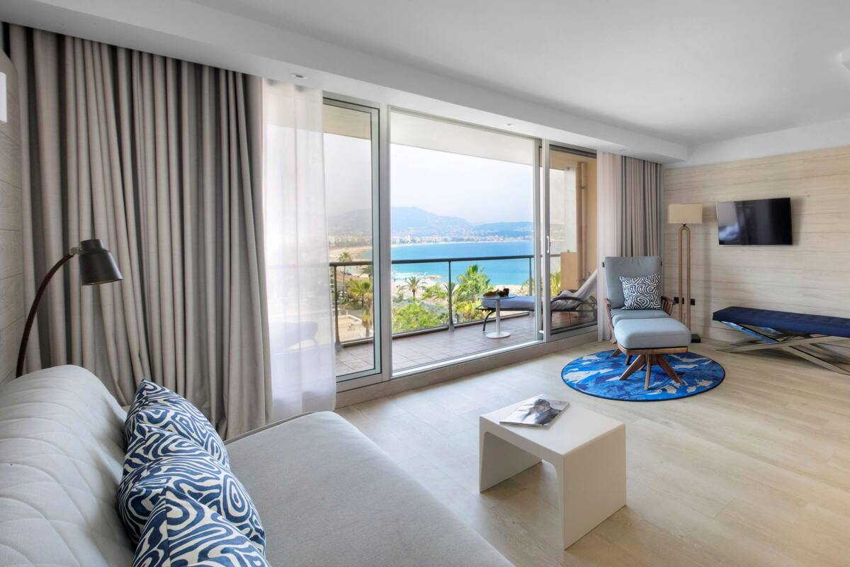 A modern hotel room with a large balcony, a sofa, a chair, and a dining area.