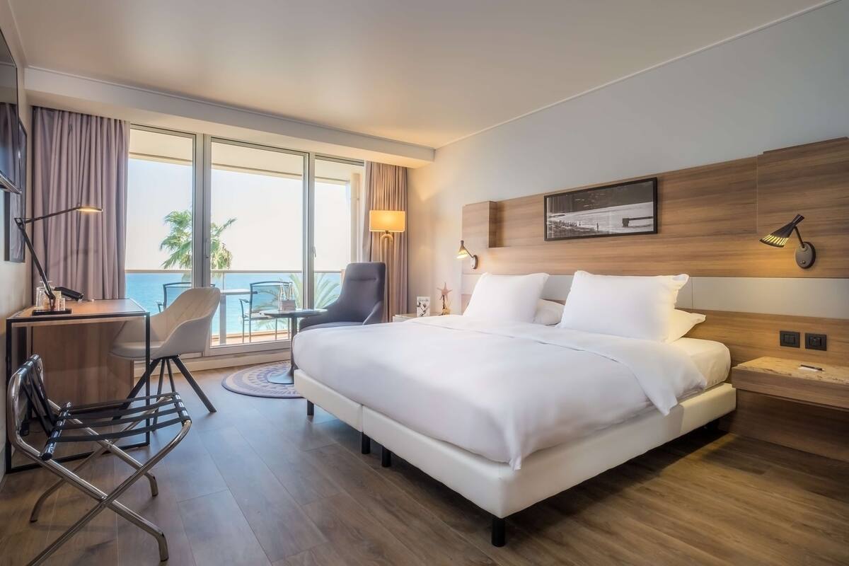 A hotel room with a large bed, a desk with a chair, a small table, and a balcony with a view of palm trees.