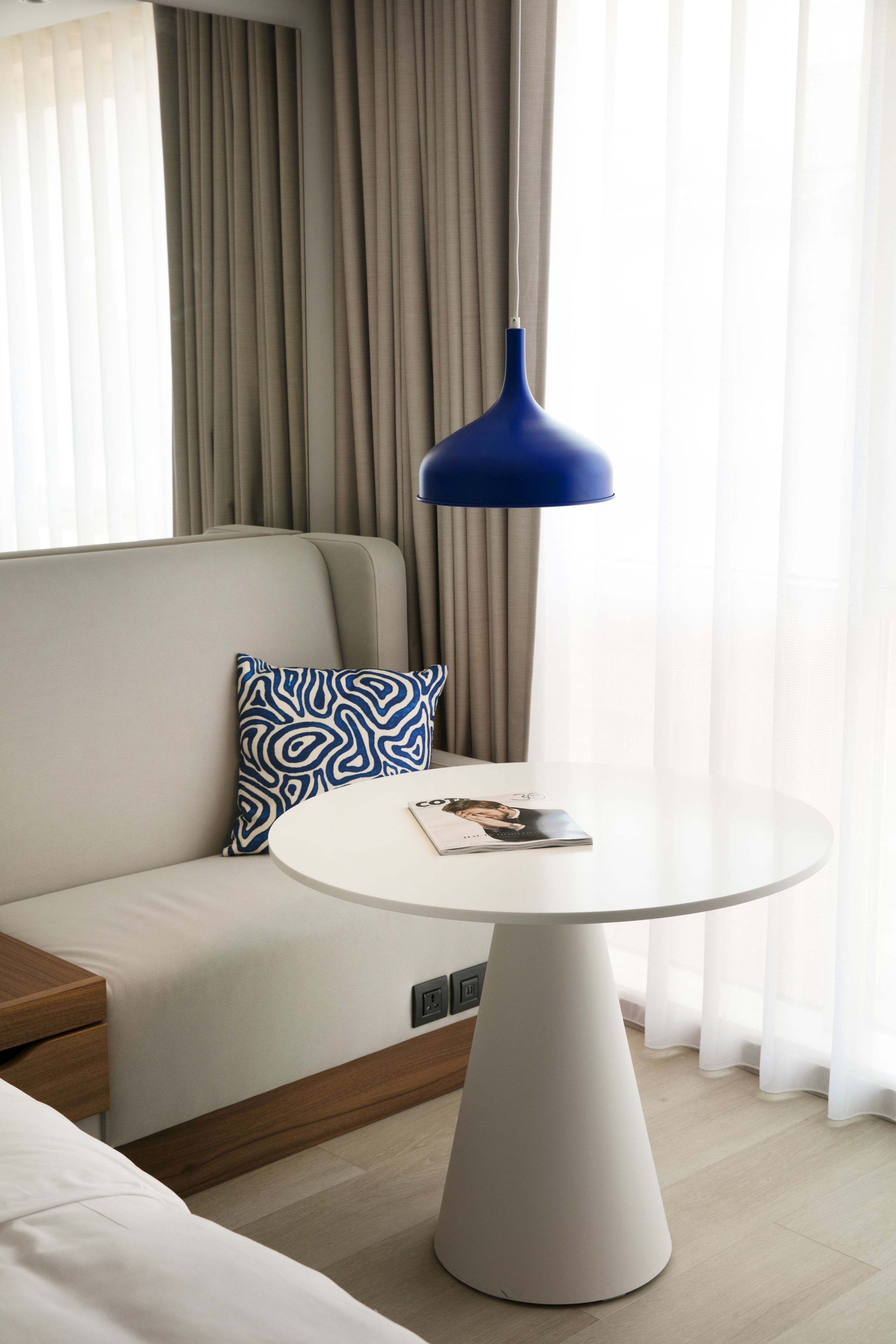 A modern hotel room with a round white table, a blue pendant lamp, a patterned pillow, and a sofa.