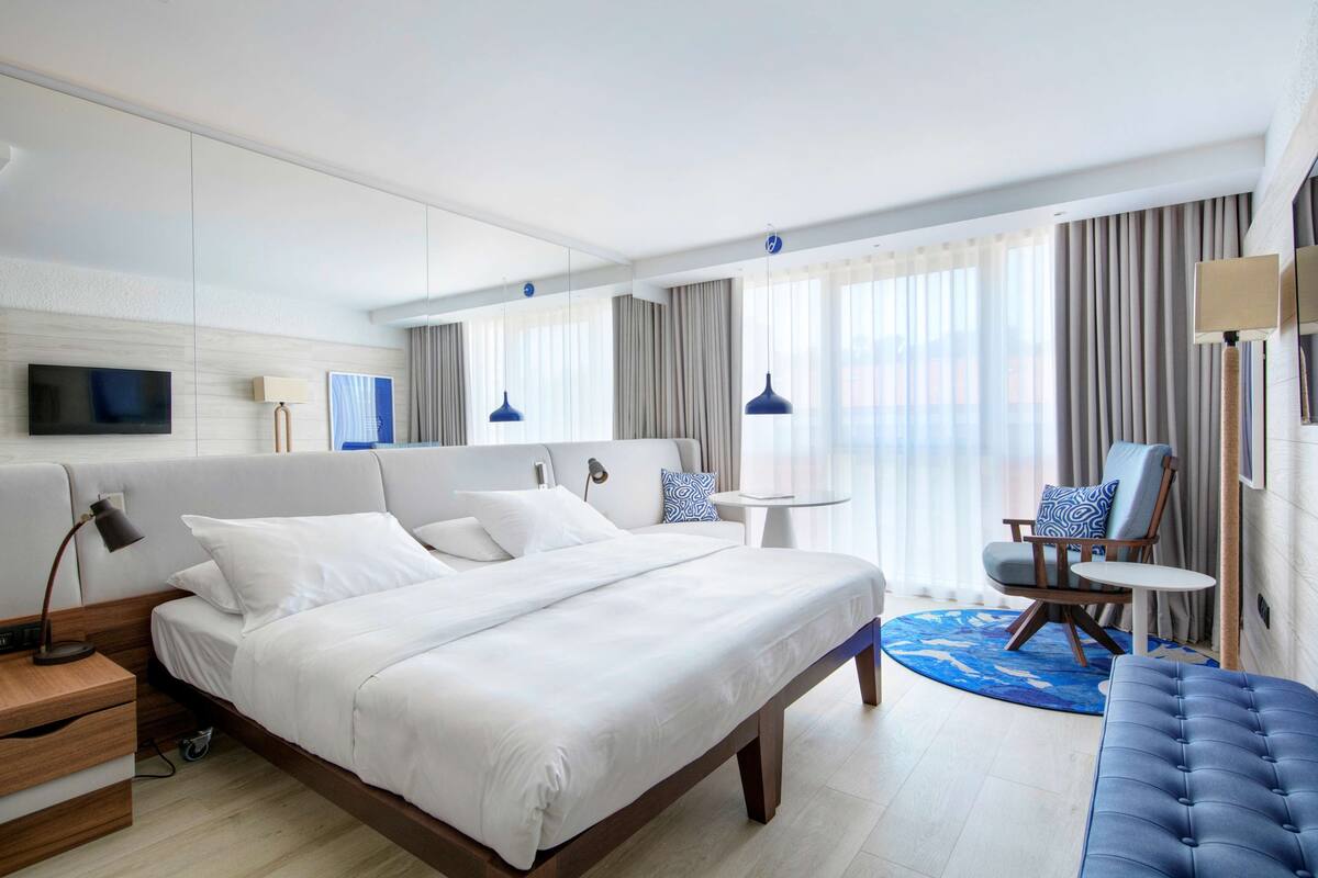 A modern hotel room with a large bed, a seating area with a chair and table, and a television mounted on the wall.