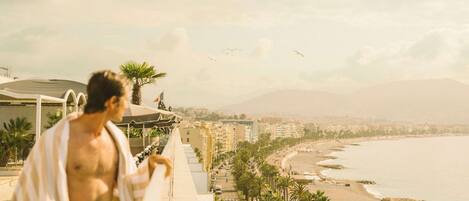 Un homme se promène sur un balcon qui donne sur une plage, avec des palmiers et des bâtiments en arrière-plan.