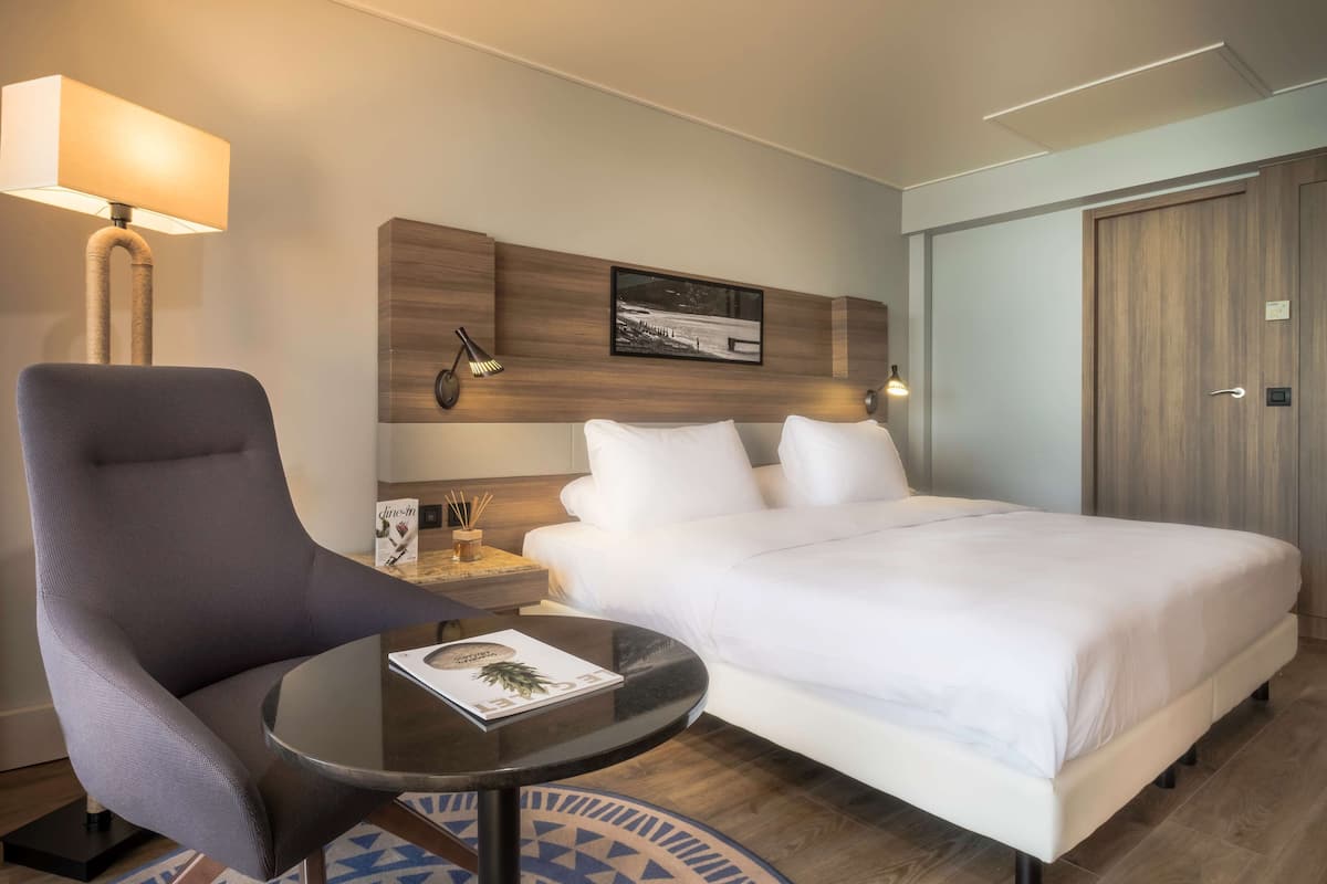 A hotel room with a large bed, a chair, a small table, and a lamp.
