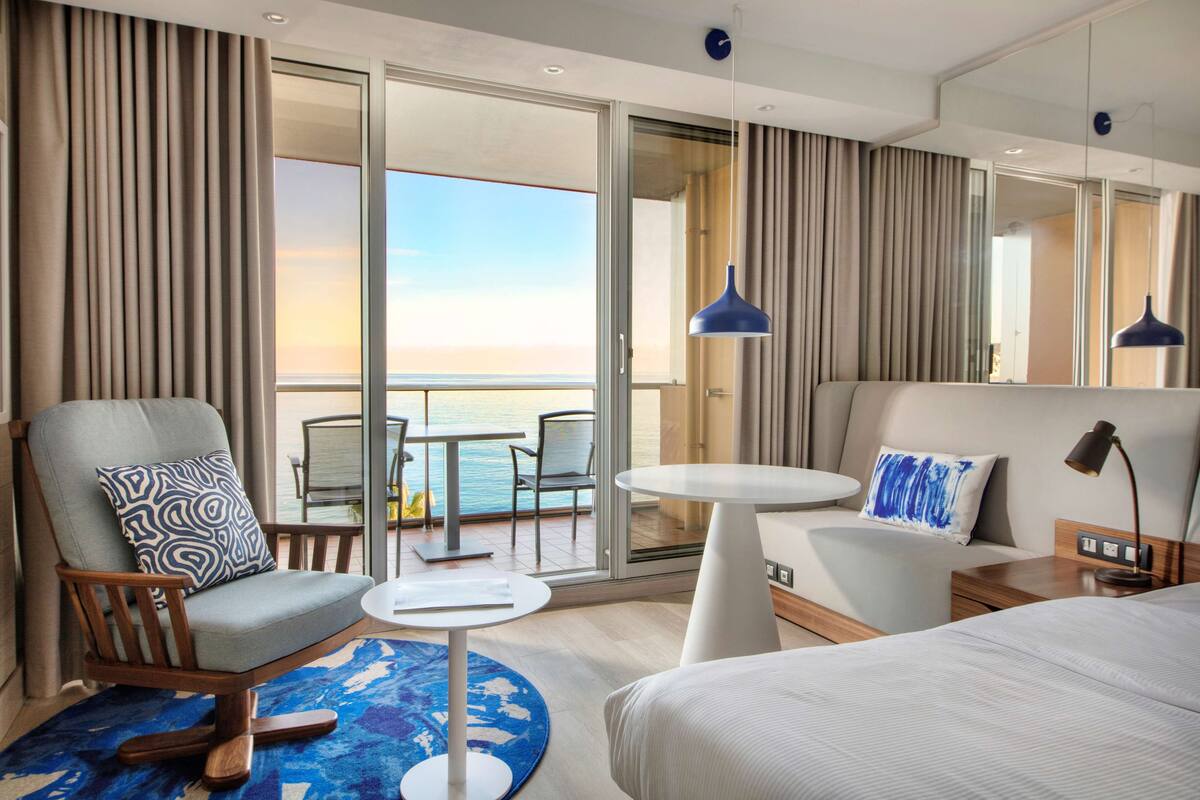 A modern hotel room with a balcony, a sofa, a chair, a bed, and a small table.