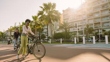 Una persona montando en bicicleta por un sendero pavimentado, con palmeras y un edificio de varios pisos al fondo.