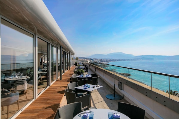 A balcony with tables and chairs, overlooking a coastal view.