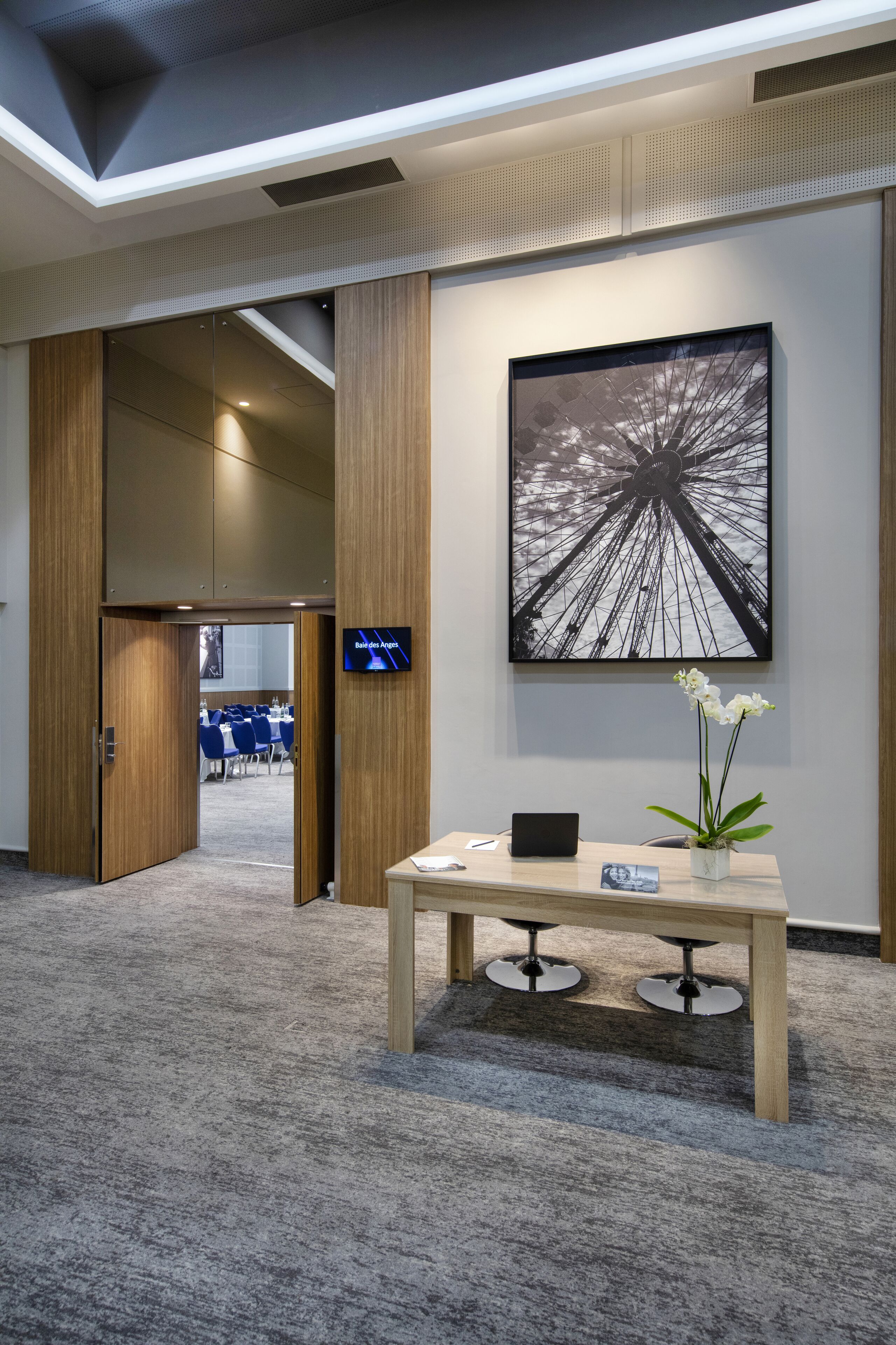 Un hall d’hôtel moderne, agrémenté d’une grande œuvre d’art représentant une grande roue, d’un comptoir de réception avec un ordinateur portable et d’une orchidée en pot.