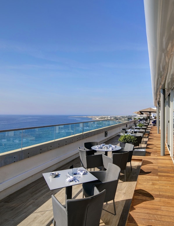A rooftop terrace with tables and chairs, overlooking a coastal view.