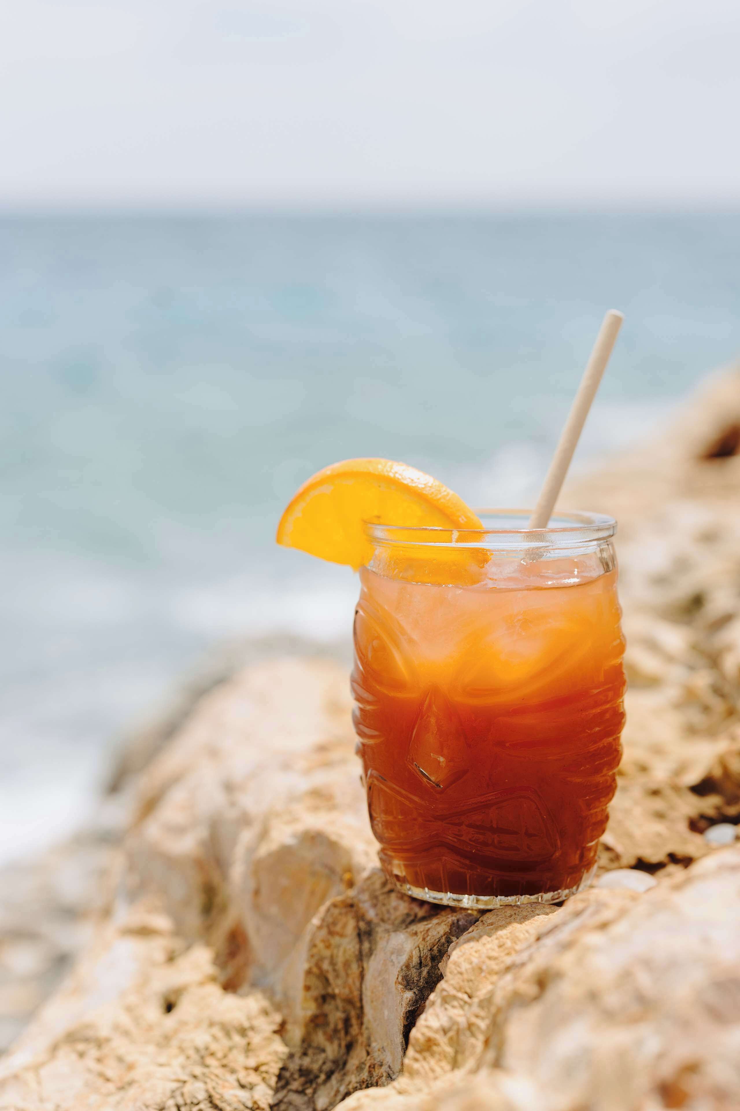 A glass of iced tea with an orange slice on a rock by the ocean.
