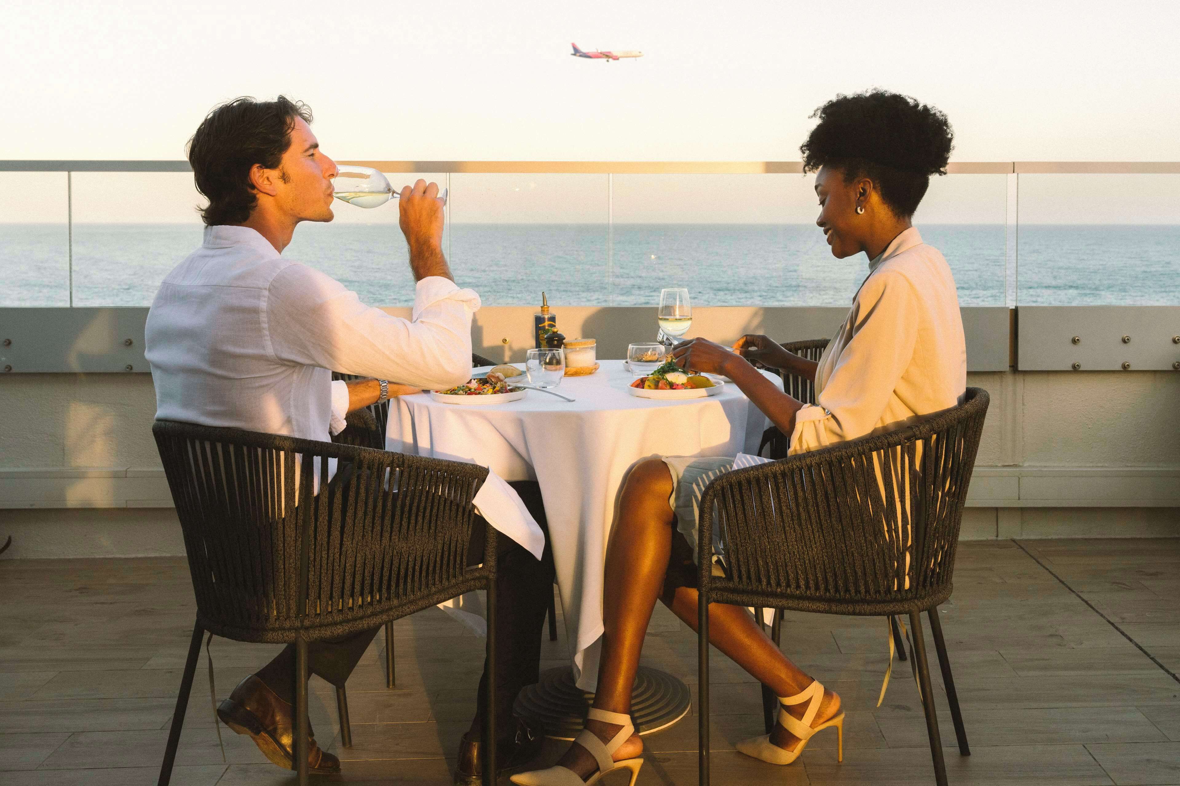 A couple dining at a table with a view of the ocean and an airplane in the distance.