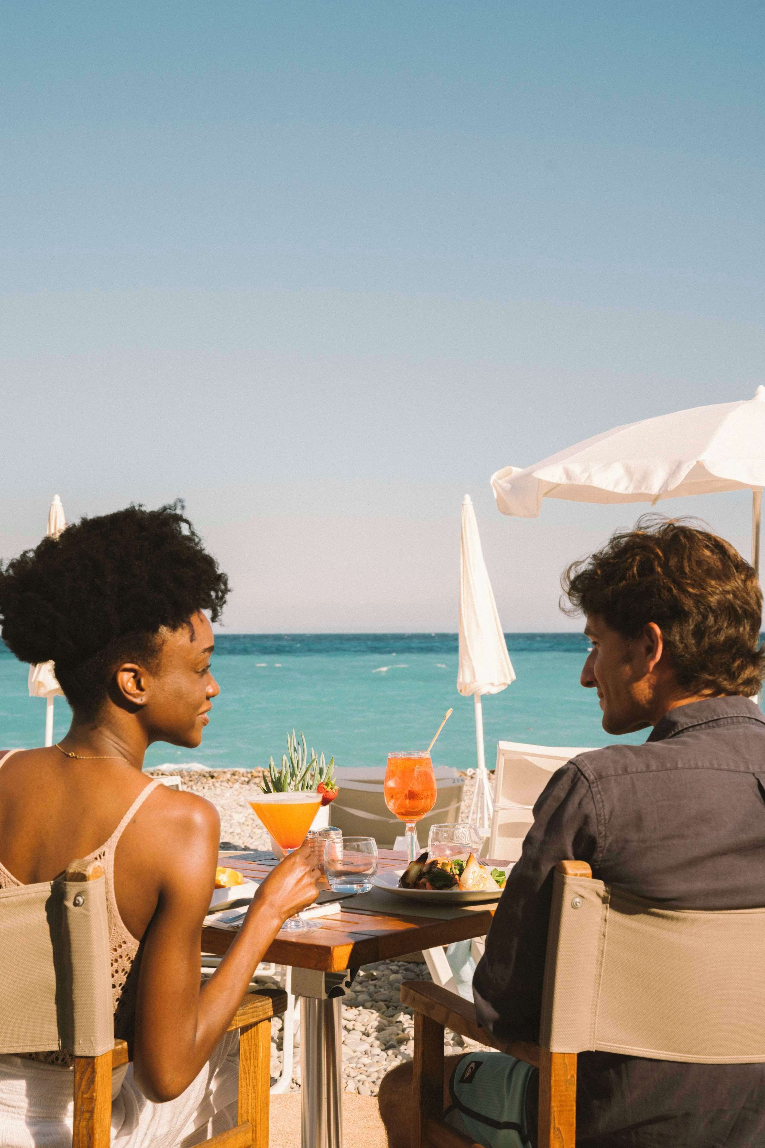 A table with a drink, a plate of food, and a potted plant at a beachside table with a white umbrella.