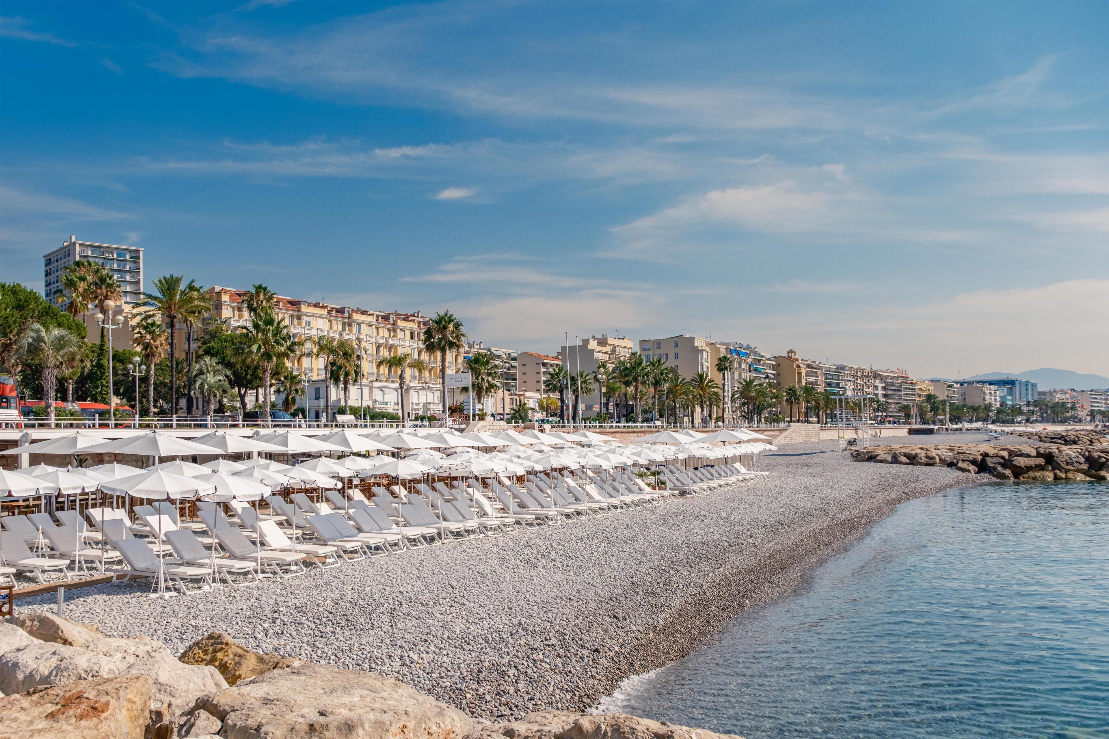 A beach with rows of sun loungers and umbrellas, a pebble beach, and buildings in the background.