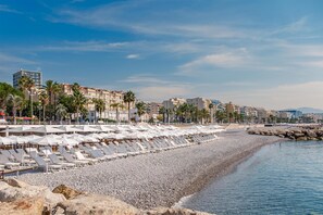 Una playa con hileras de tumbonas y sombrillas, arena de guijarros y edificios al fondo.