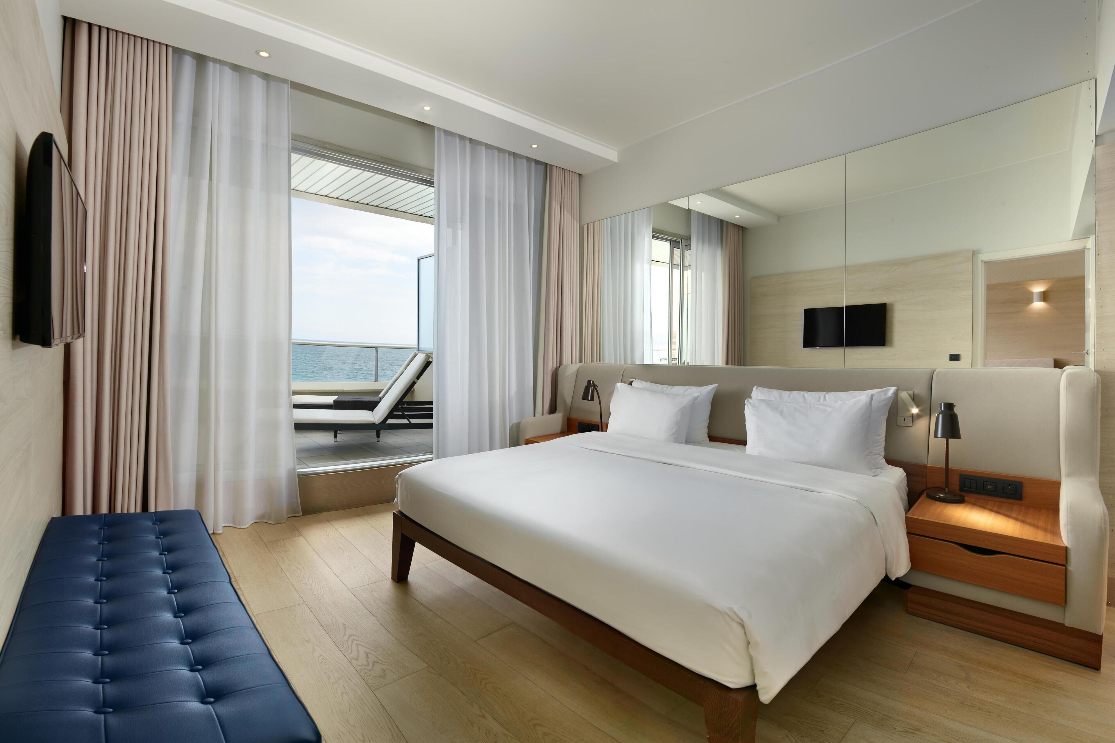 A modern hotel room with a large bed, a bench, a TV, and a balcony with a view of the sea.