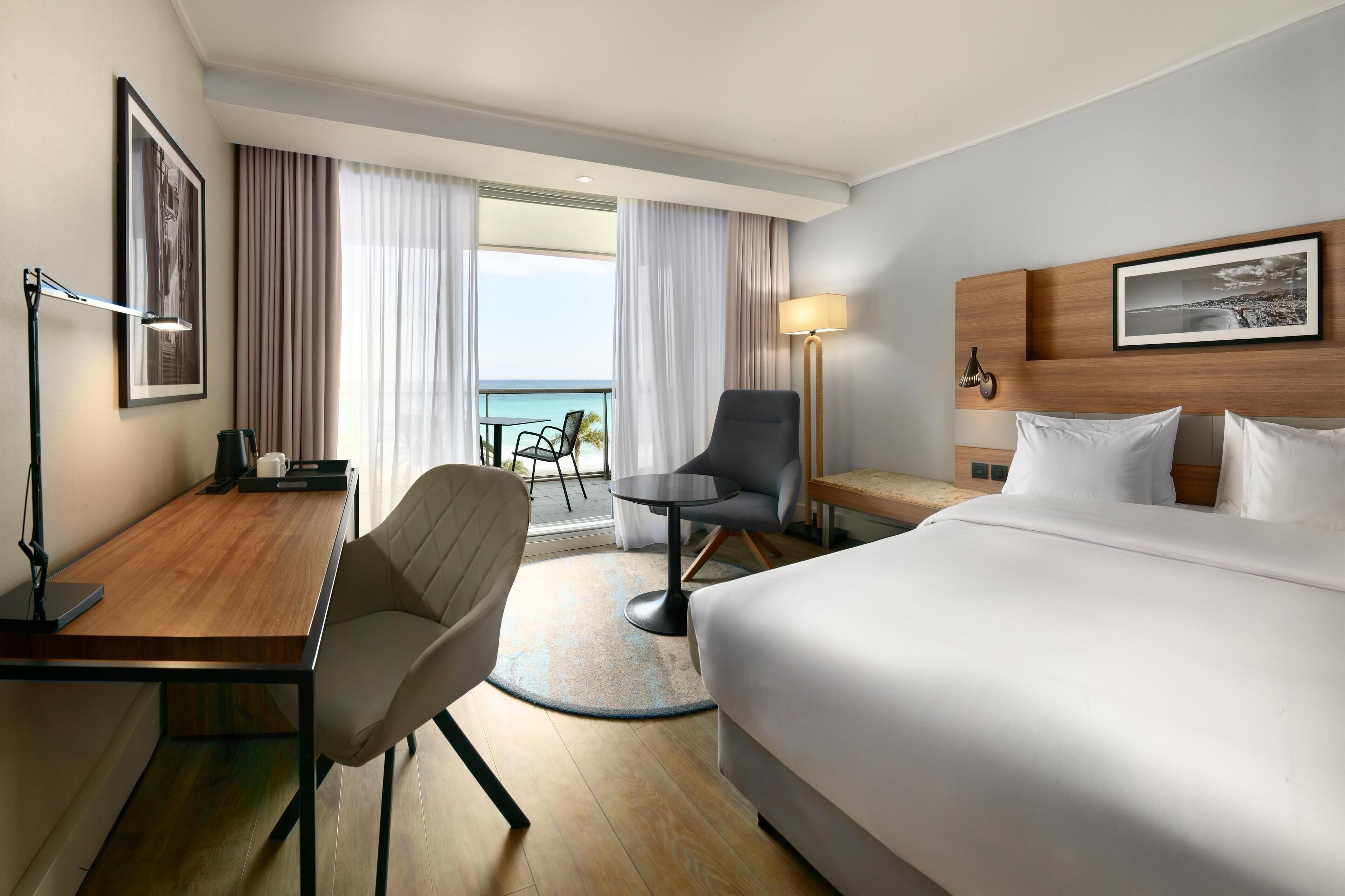 A hotel room with a bed, a desk with a chair, a round table with a chair, a lamp, and a view of the sea through large windows.