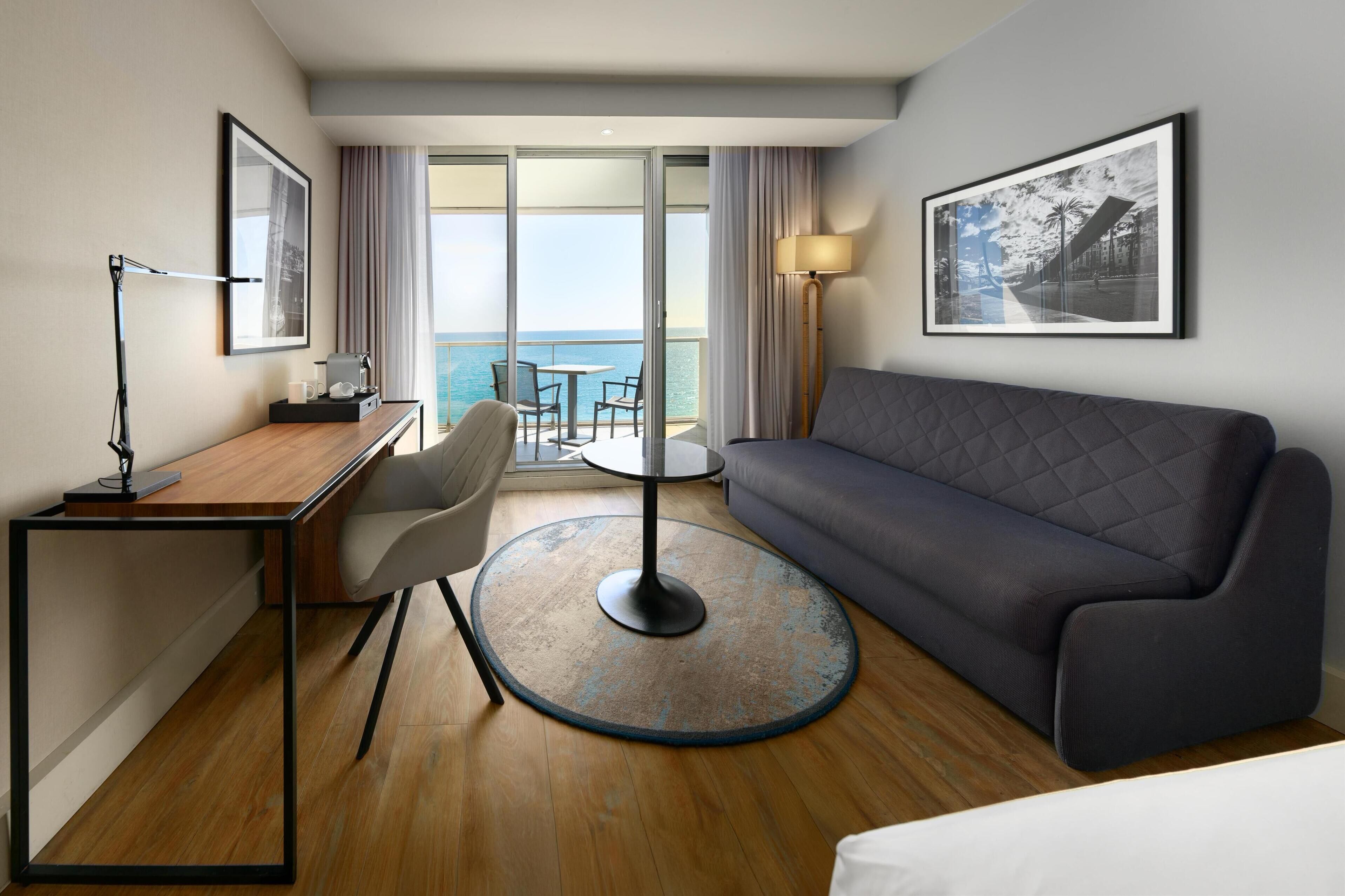 A hotel room with a bed, a desk with a chair, a round table, a sofa, a large window with a view of the sea, and two framed pictures on the walls.