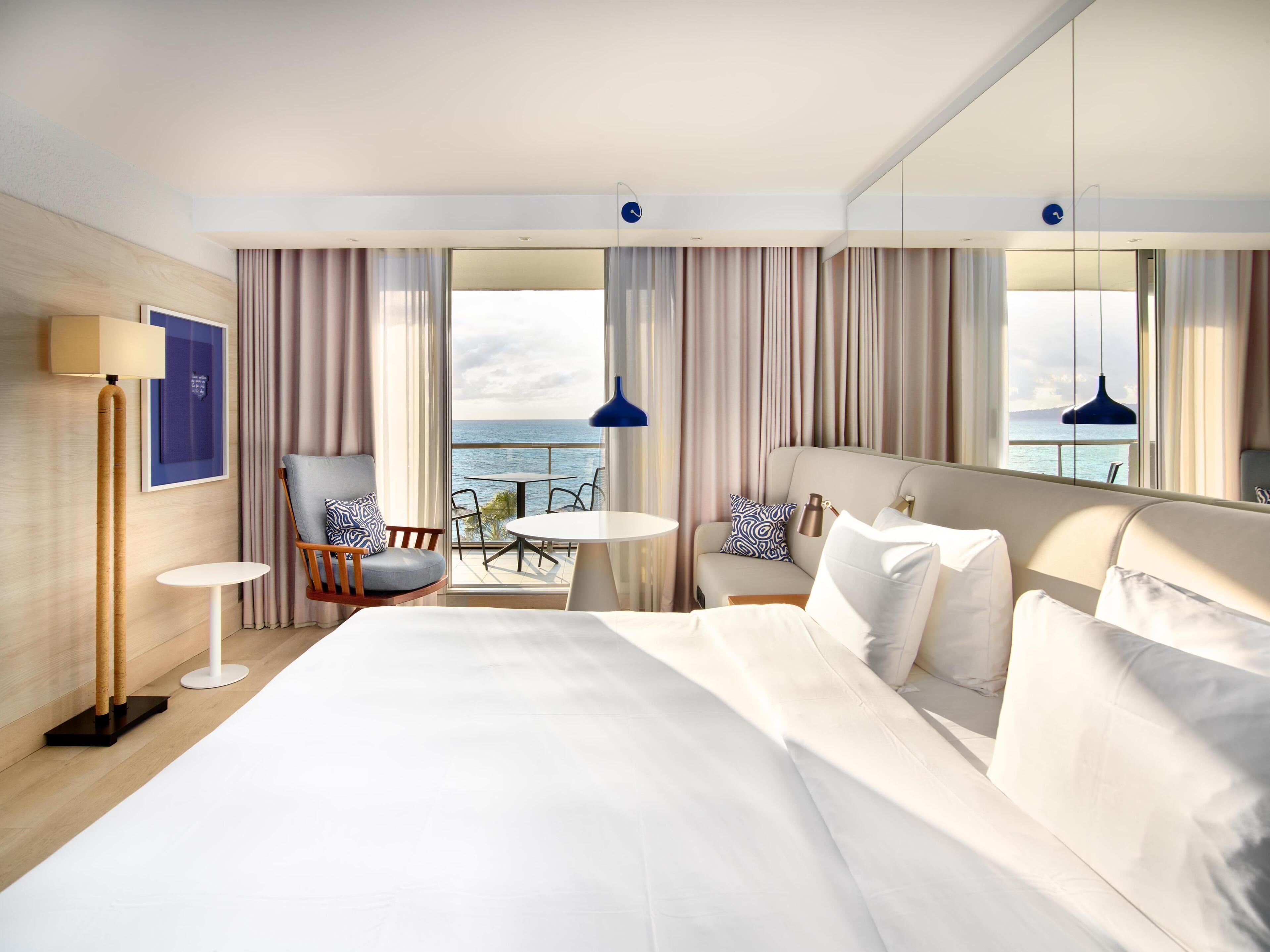 Una habitación de hotel moderna con una cama grande, una zona de estar con una mesa y sillas, un ventanal con vistas al mar y un espejo que refleja la habitación.