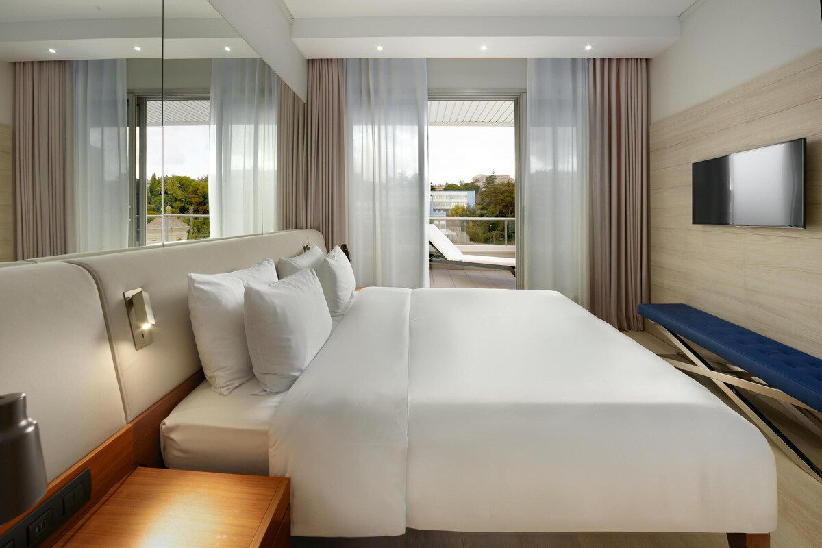 Una habitación de hotel moderna con una cama grande, un televisor de pantalla plana en la pared y vistas a la ciudad a través de amplios ventanales.