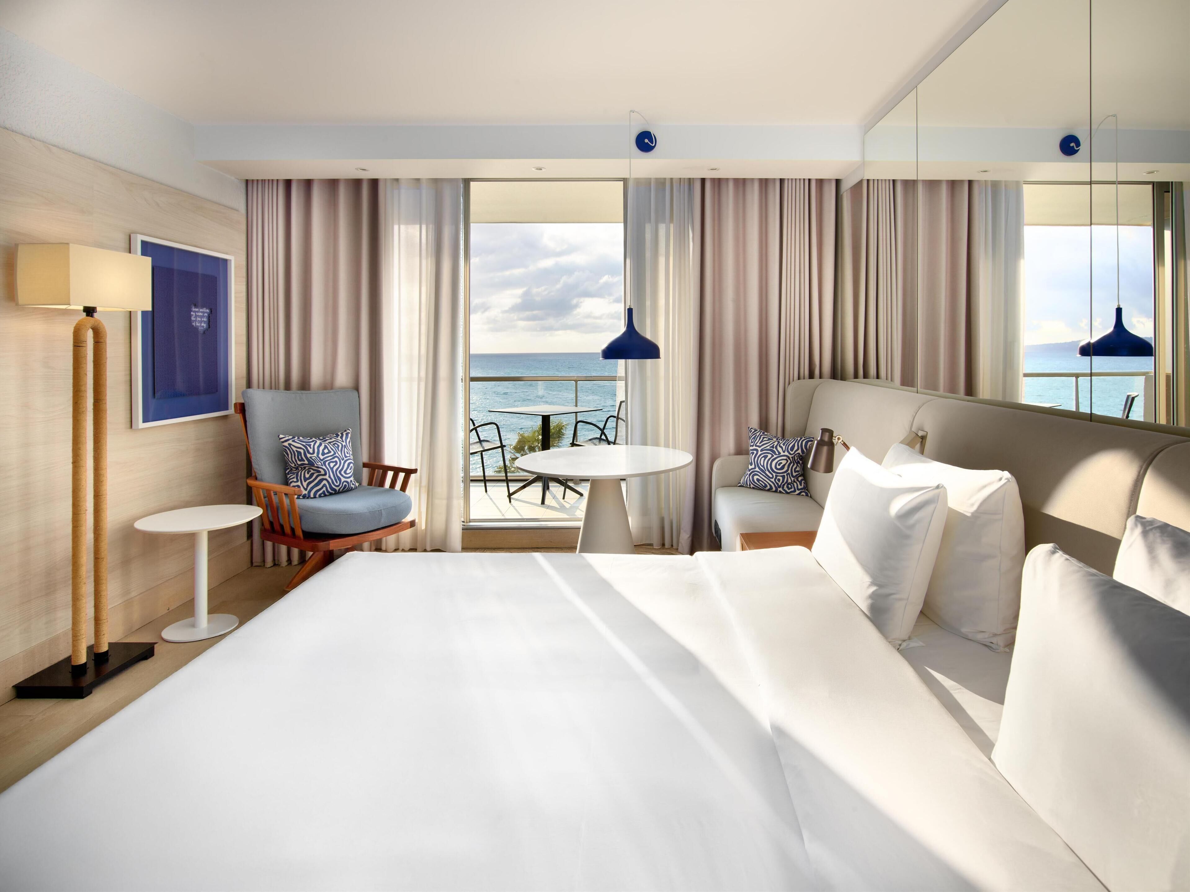 Een hotelkamer met een groot bed, een zithoek met een stoel en tafel, en een balkon met uitzicht op zee.