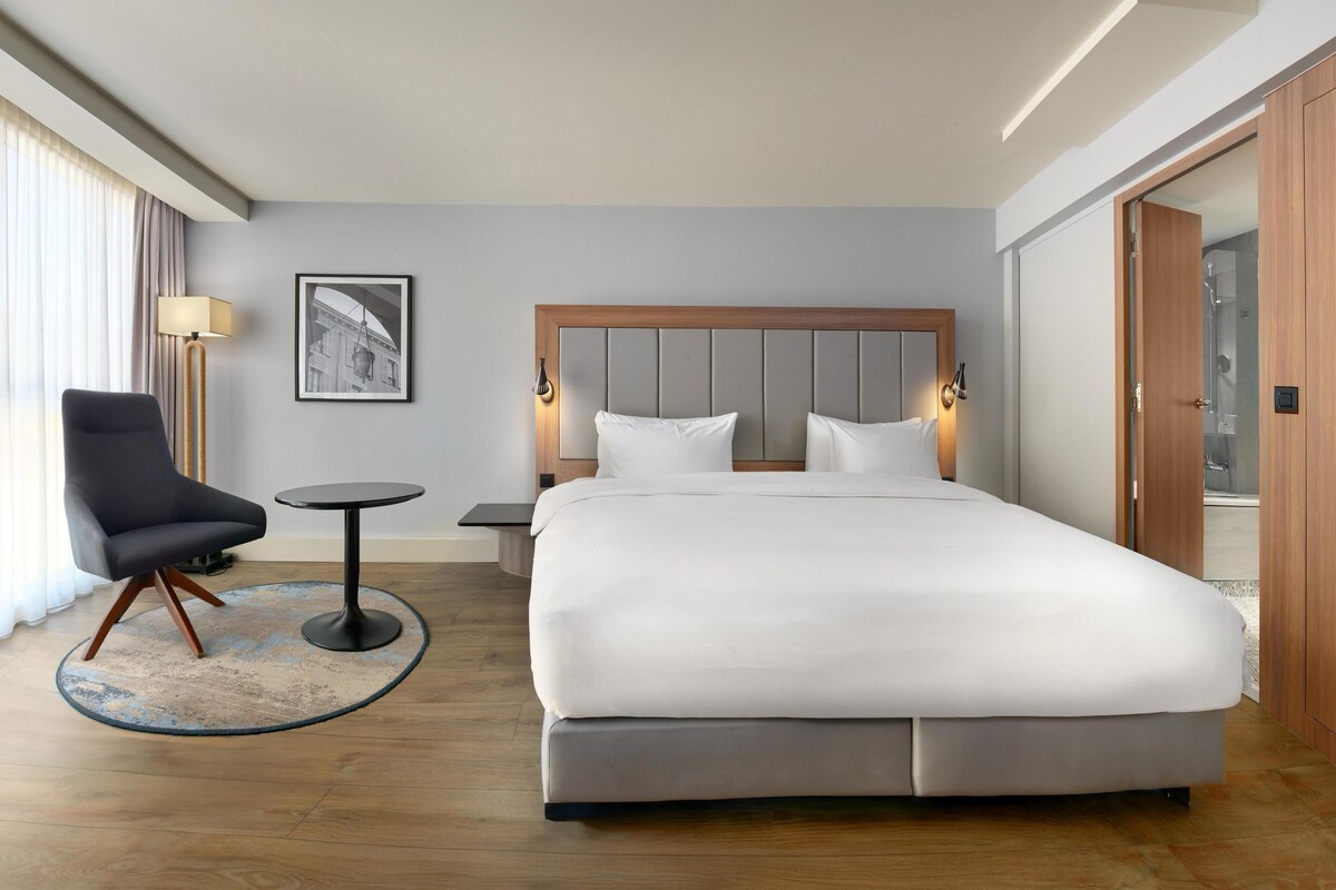Ein Hotelzimmer mit einem großen Bett, einem Sessel, einem kleinen Tisch und einem Bild an der Wand.
