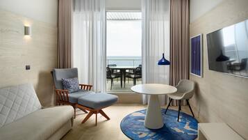 Ett modernt hotellrum med en soffa, två fåtöljer, ett runt bord och utsikt över havet genom stora fönster.