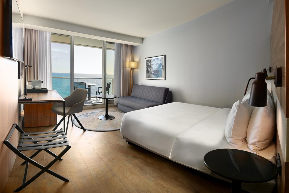 Habitación de hotel con cama, escritorio, silla, sofá, mesita, lámpara y ventana con vista al océano.