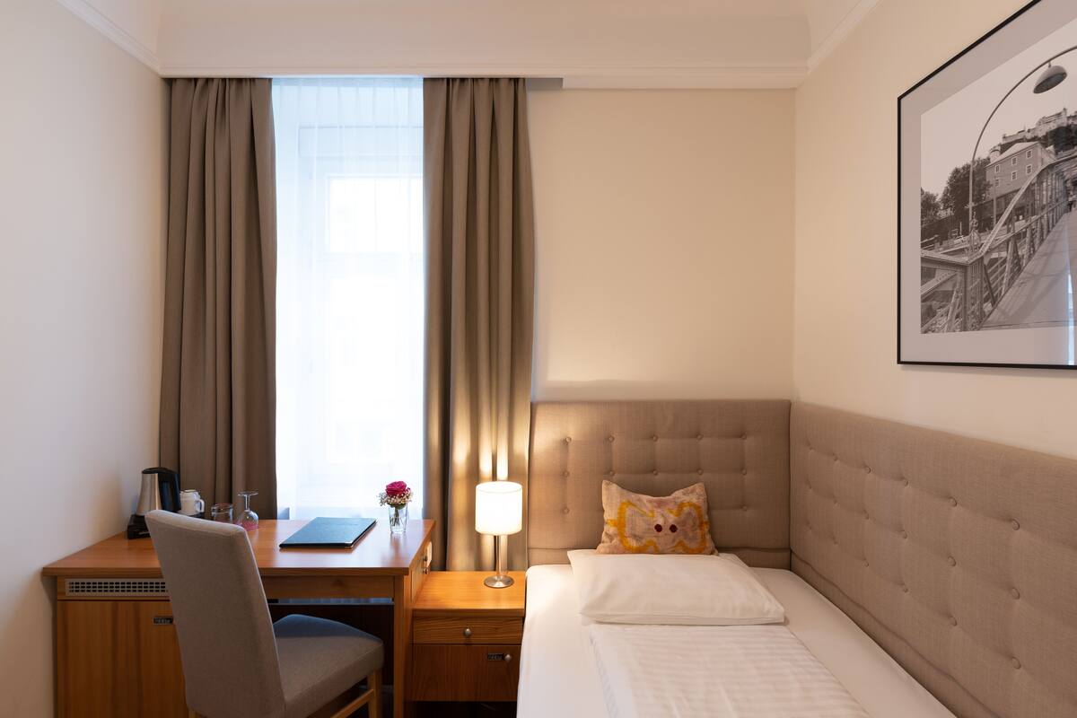 A hotel room with a bed, a desk, a chair, a lamp, a nightstand, and a framed picture on the wall.