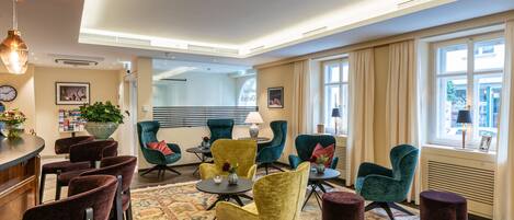 A hotel lobby with a bar area, seating with a patterned rug, and large windows with curtains.