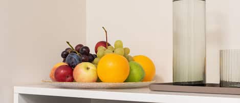 Estante blanco con un frutero que contiene manzanas, naranjas, uvas y un limón, un jarrón alto de vidrio y toallas dobladas con cuidado.