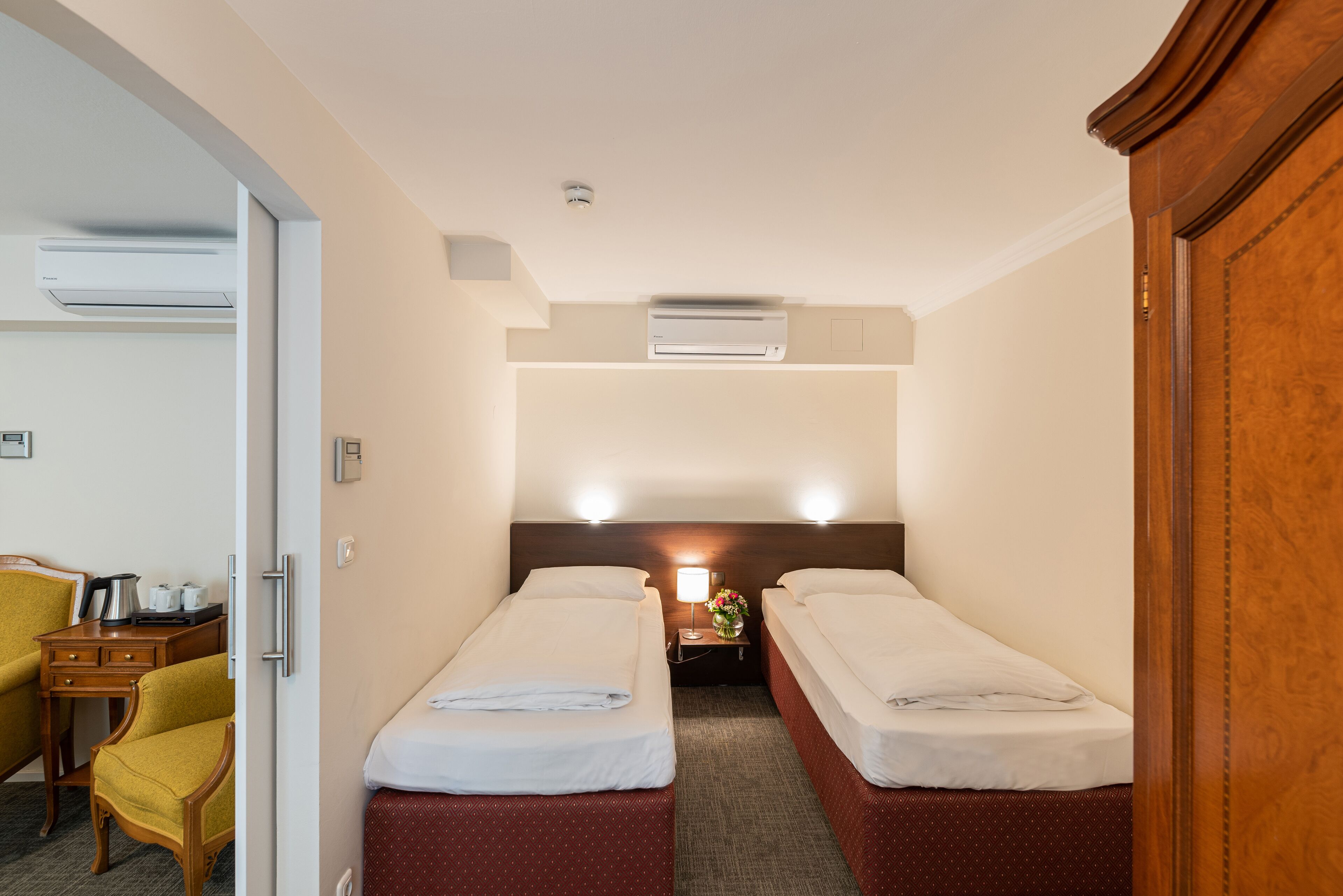 Ein Hotelzimmer mit zwei Einzelbetten, einem Schreibtisch mit Stuhl, einer Klimaanlage und einem Nachttisch mit Lampe und Blumen.
