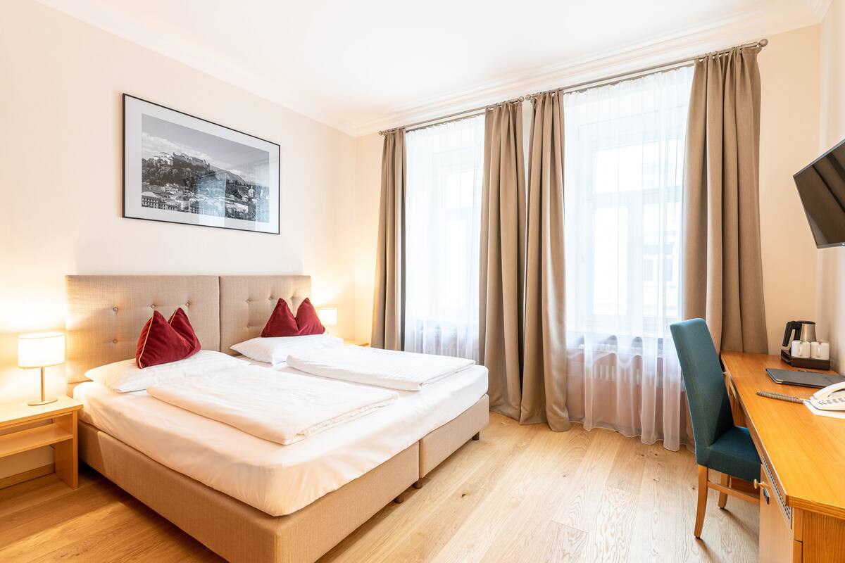 A hotel room with a bed, a desk, a chair, a TV, and a window with curtains.