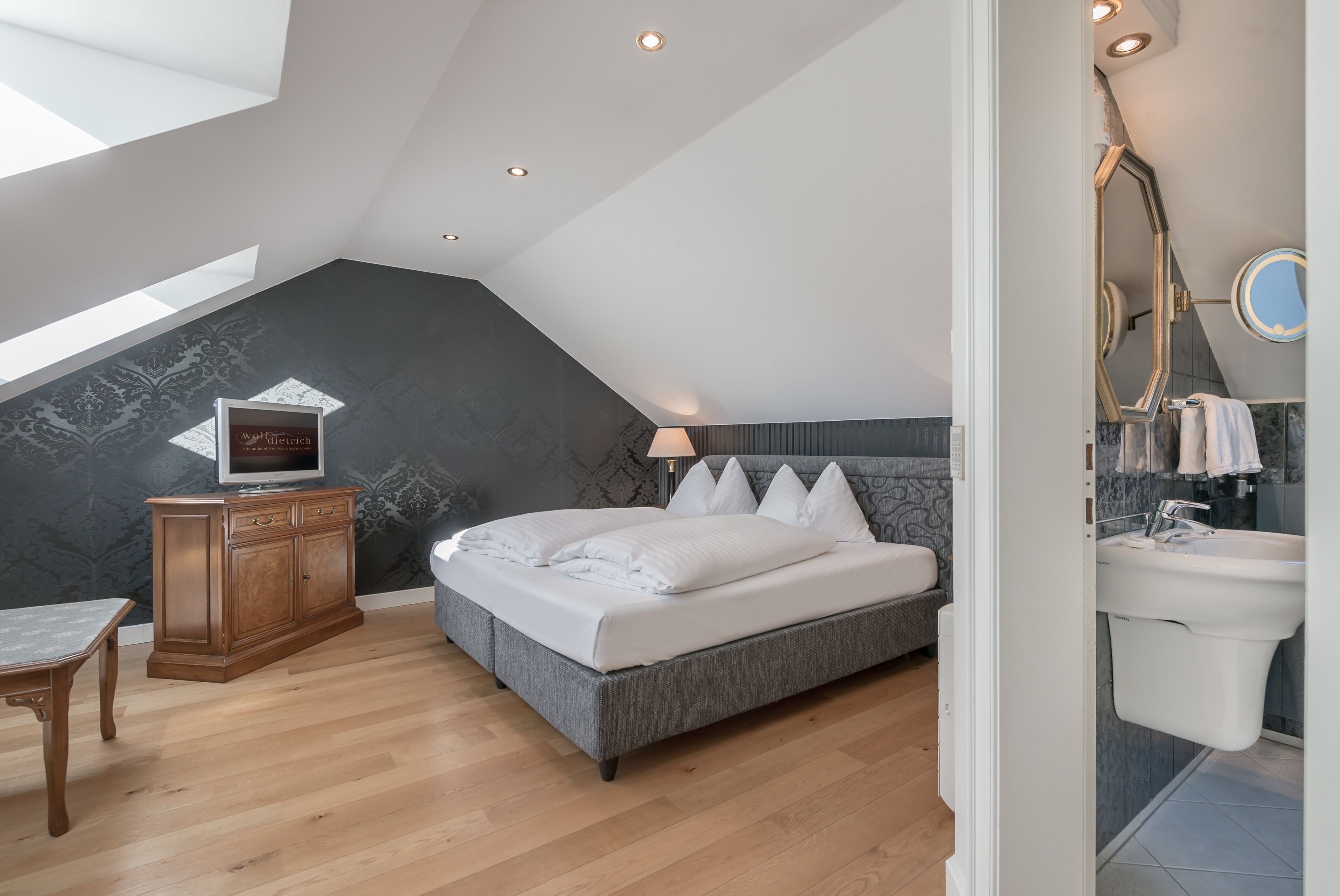 A bedroom with a sloped ceiling, a bed, a wooden cabinet, a TV, and a bathroom with a sink and mirror.