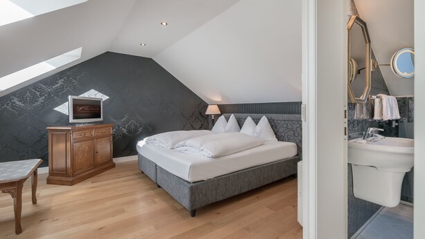 A bedroom with a sloped ceiling, a bed, a wooden cabinet, a TV, and a bathroom with a sink and mirror. - Altstadthotel Wolf-Dietrich (Salzburg)