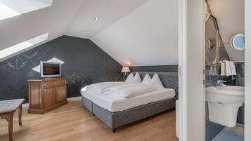 A bedroom with a sloped ceiling, a bed, a wooden cabinet, a TV, and a bathroom with a sink and mirror.