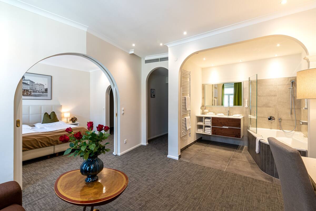 A modern hotel room with a bed, a sitting area with a vase of flowers, a bathroom with a bathtub and shower, and a hallway with an archway.