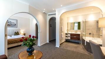 A modern hotel room with a bed, a sitting area with a vase of flowers, a bathroom with a bathtub and shower, and a hallway with an archway.