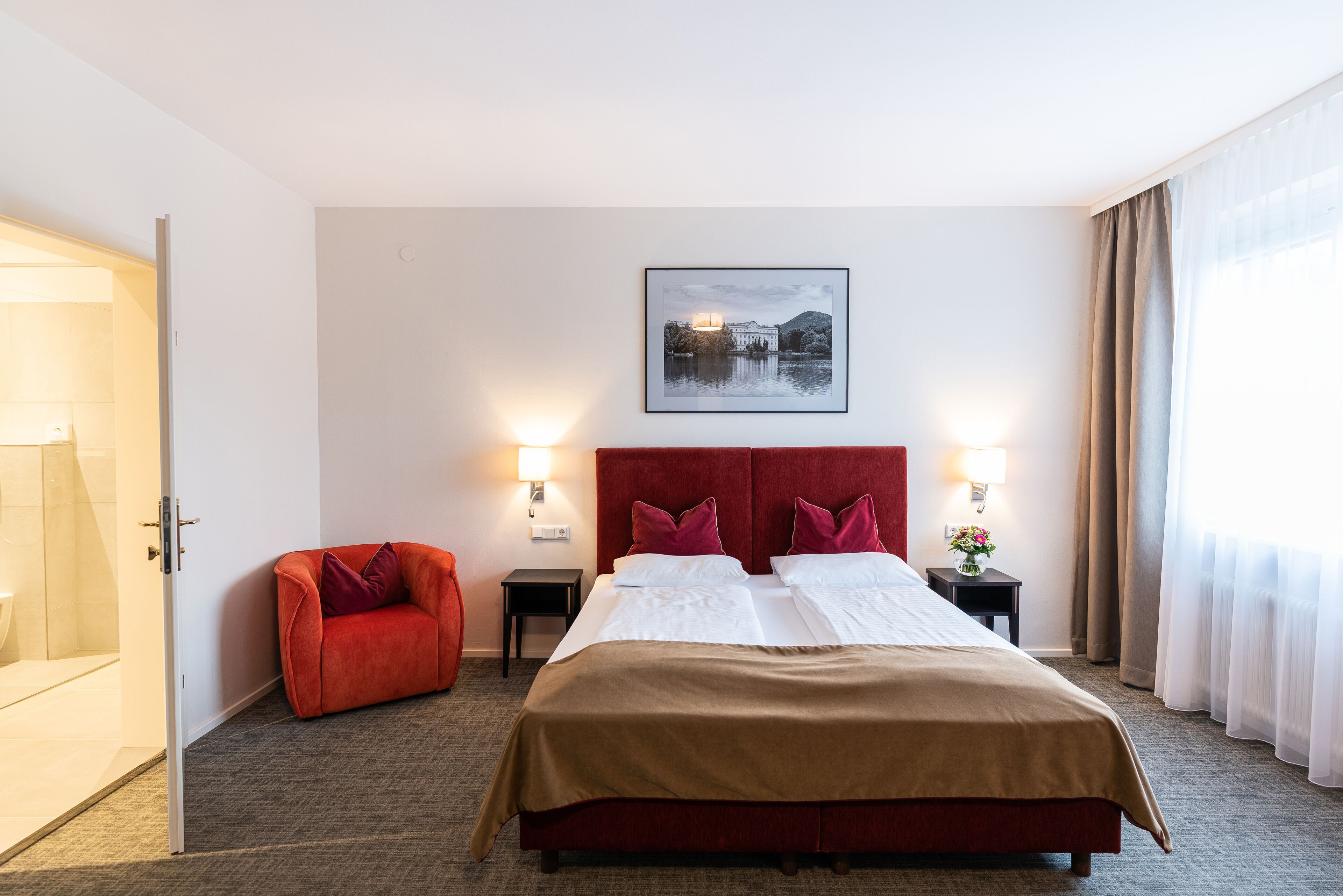 Ein Hotelzimmer mit einem großen Bett, zwei Nachttischen, einem roten Sessel, einem gerahmten Bild an der Wand und einer Badezimmertür.