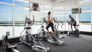 A gym with treadmills and elliptical machines, large windows with a view of the beach, and a mounted TV. - Hotel Cardoso (Maputo)