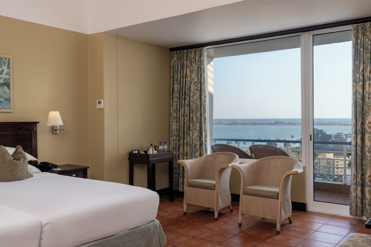 A hotel room with a bed, a desk, two wicker chairs, a view of the sea, and a painting on the wall.