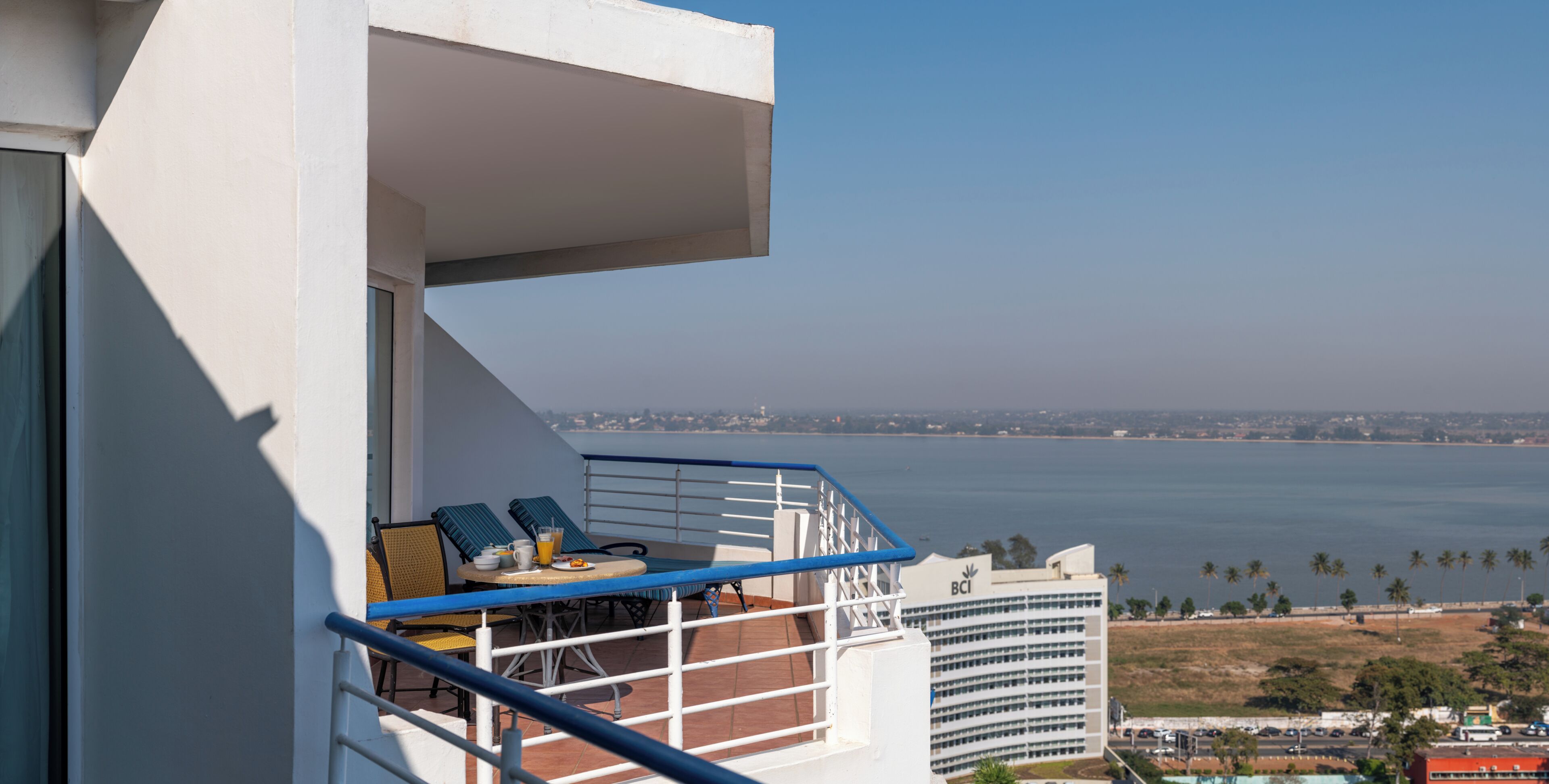 A balcony with a table, chairs, and a view of the sea and buildings.