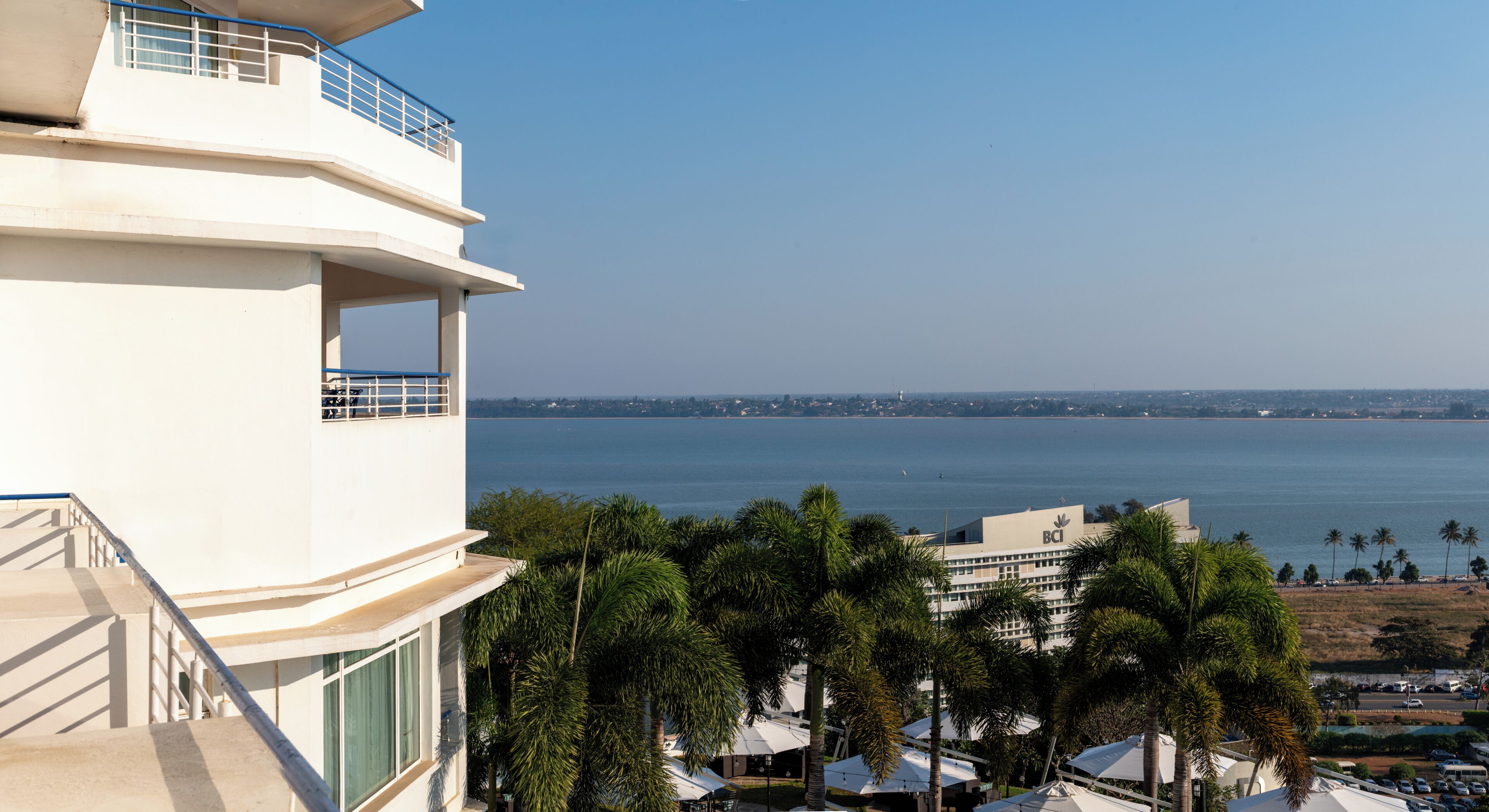 Un edificio de varios pisos con balcones que dan a una masa de agua, palmeras y una costa distante.