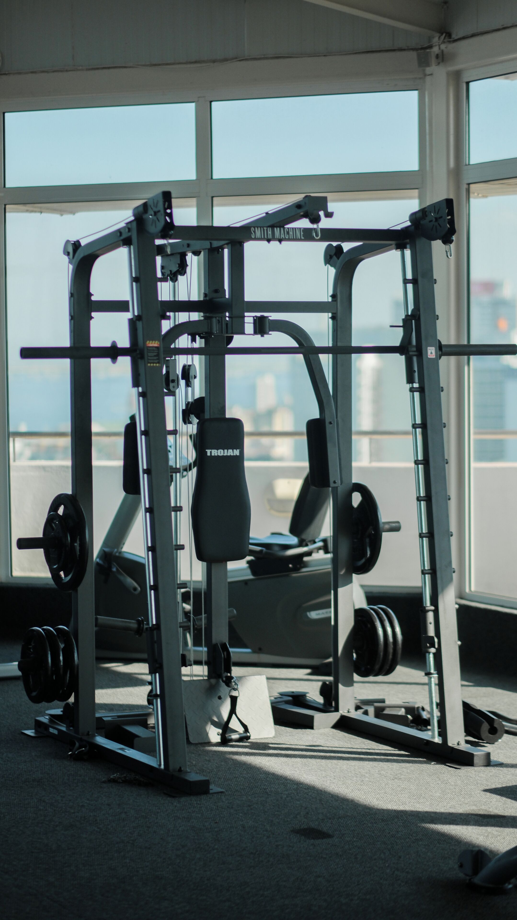 A gym with Smith Machine and TROJAN equipment, large windows, and a view of buildings.