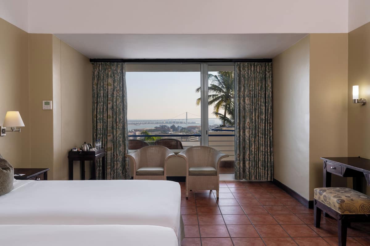 A hotel room with a bed, two chairs, a small table, a desk, and a view of a bridge and palm trees.