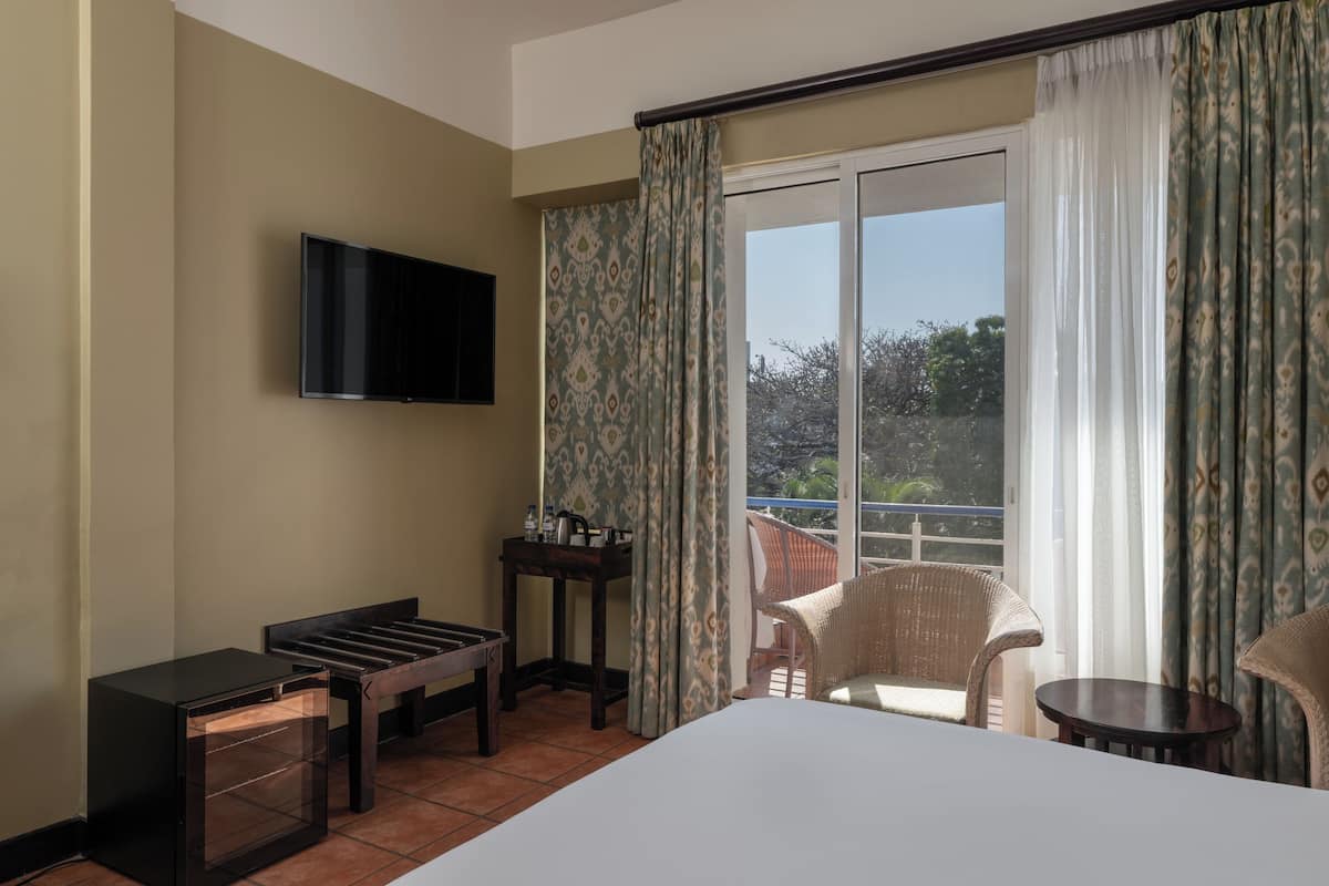A hotel room with a bed, a TV mounted on the wall, a small table with a kettle, a chair, and a balcony with curtains.