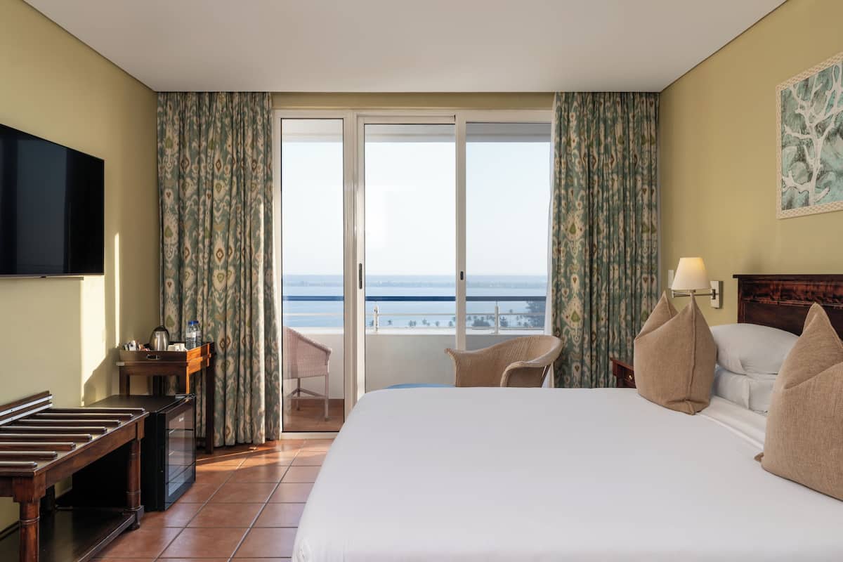 A hotel room with a bed, a desk, a TV, a view of the sea, and a balcony.