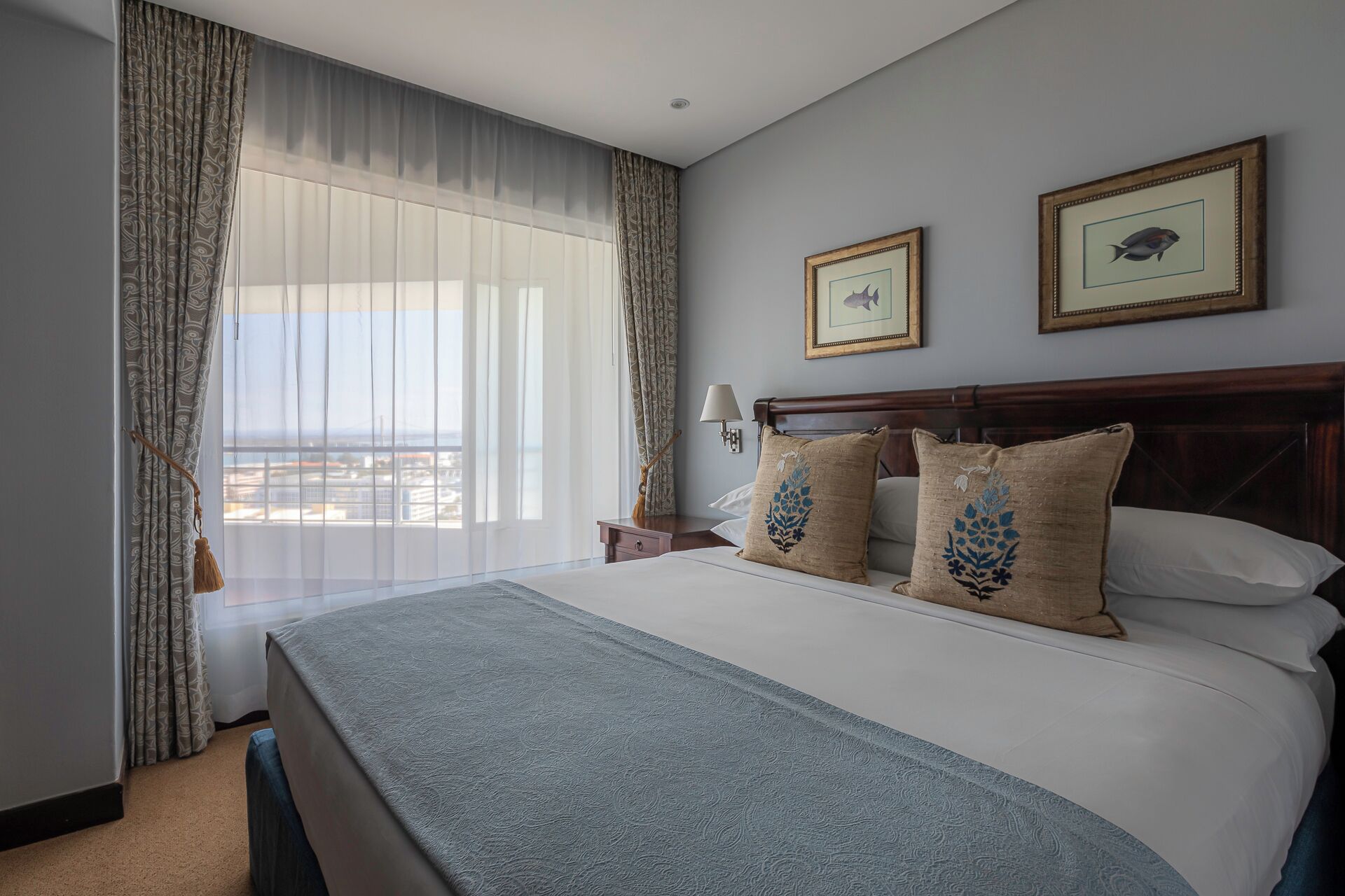 A hotel room with a large bed, two framed pictures on the wall, a nightstand, and a view of the city through the window.