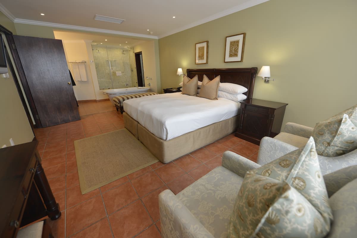 A hotel room with a large bed, a bedside table, a nightstand, a chair, a bathroom with a shower, and a door leading to another room.
