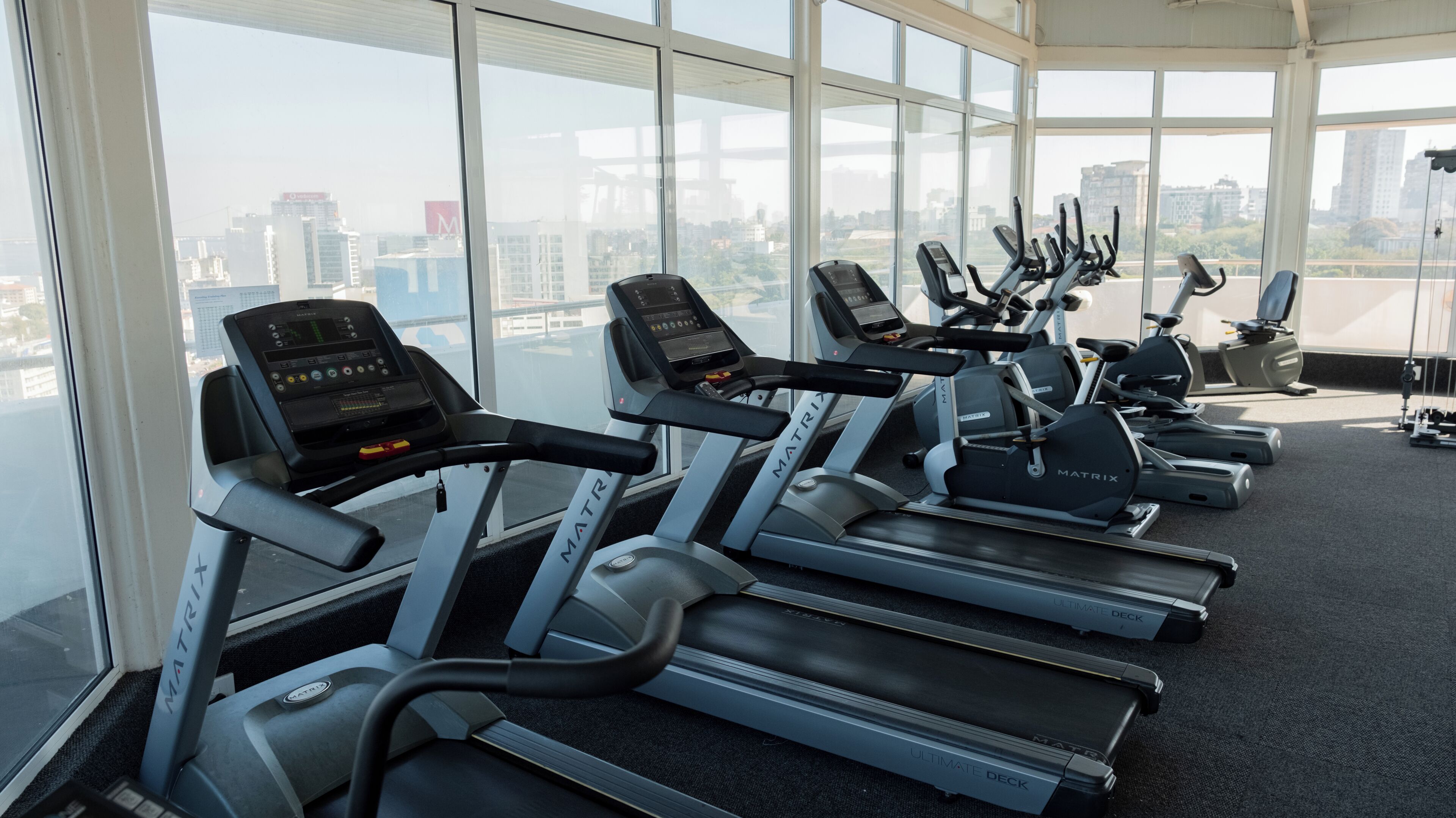 Un gimnasio con cintas de correr y elípticas, ventanas amplias y vista a la ciudad.