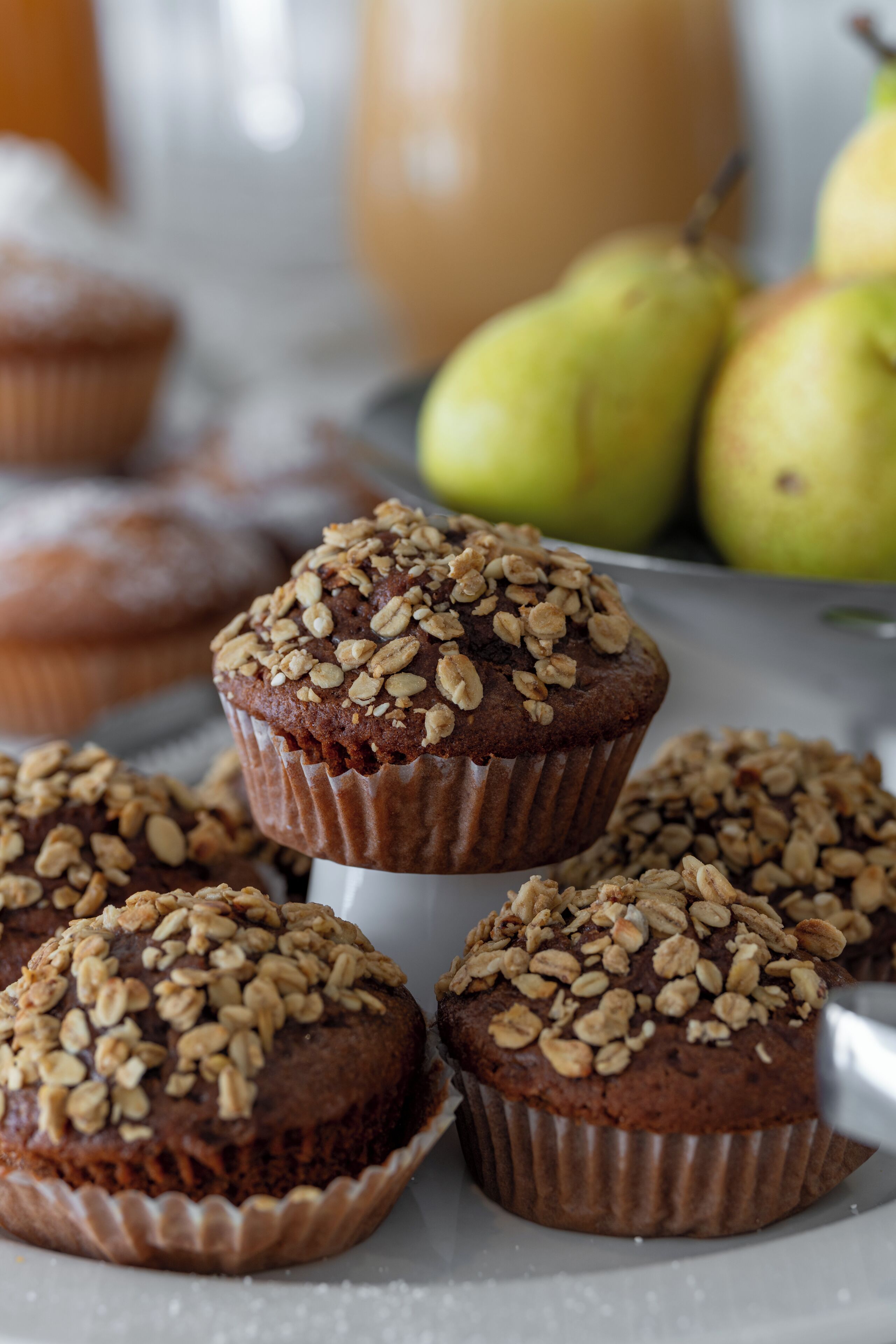 A tray of muffins topped with oats, with pears in the background.