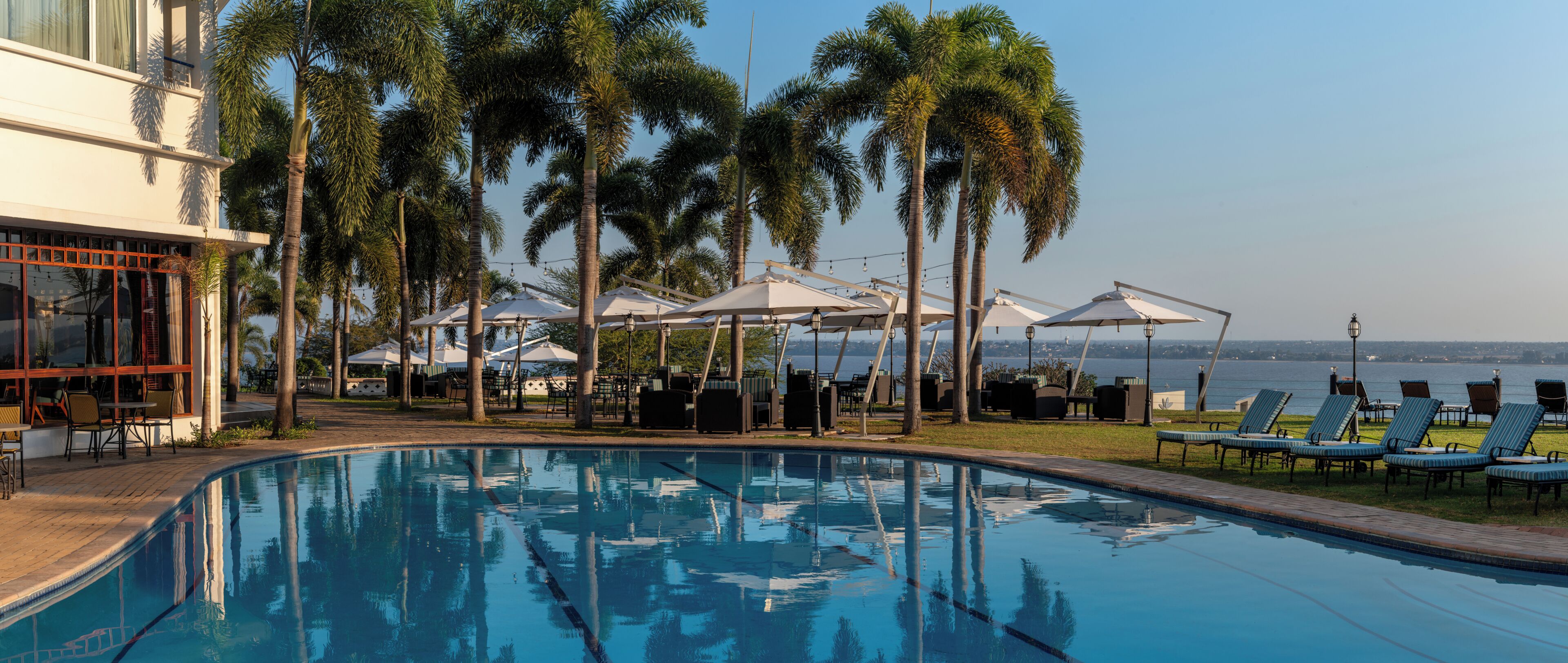Ein Swimmingpool mit Liegestühlen und Sonnenschirmen, umgeben von Palmen und mit Blick auf ein Gewässer.